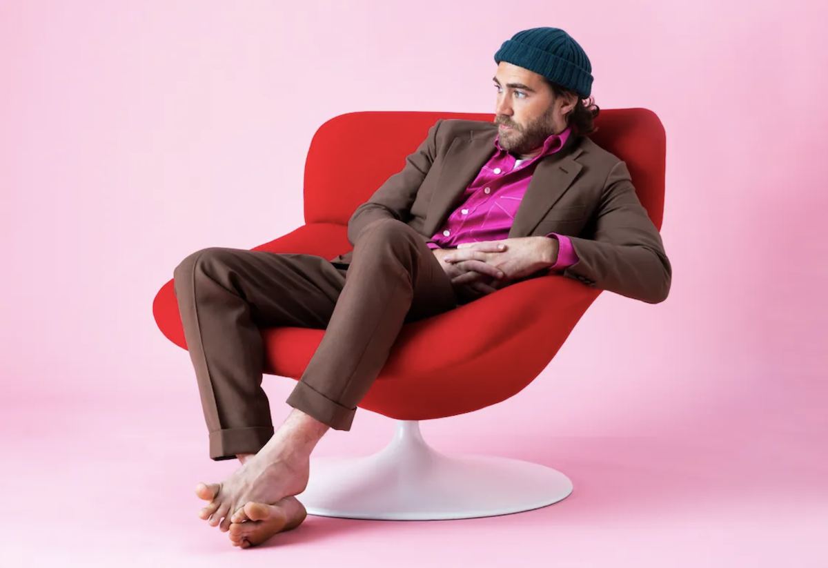 Australian musician Matt Corby sitting barefoot in a red chair wearing a brown suit and beanie for a studio promo artist portrait photo shoot.